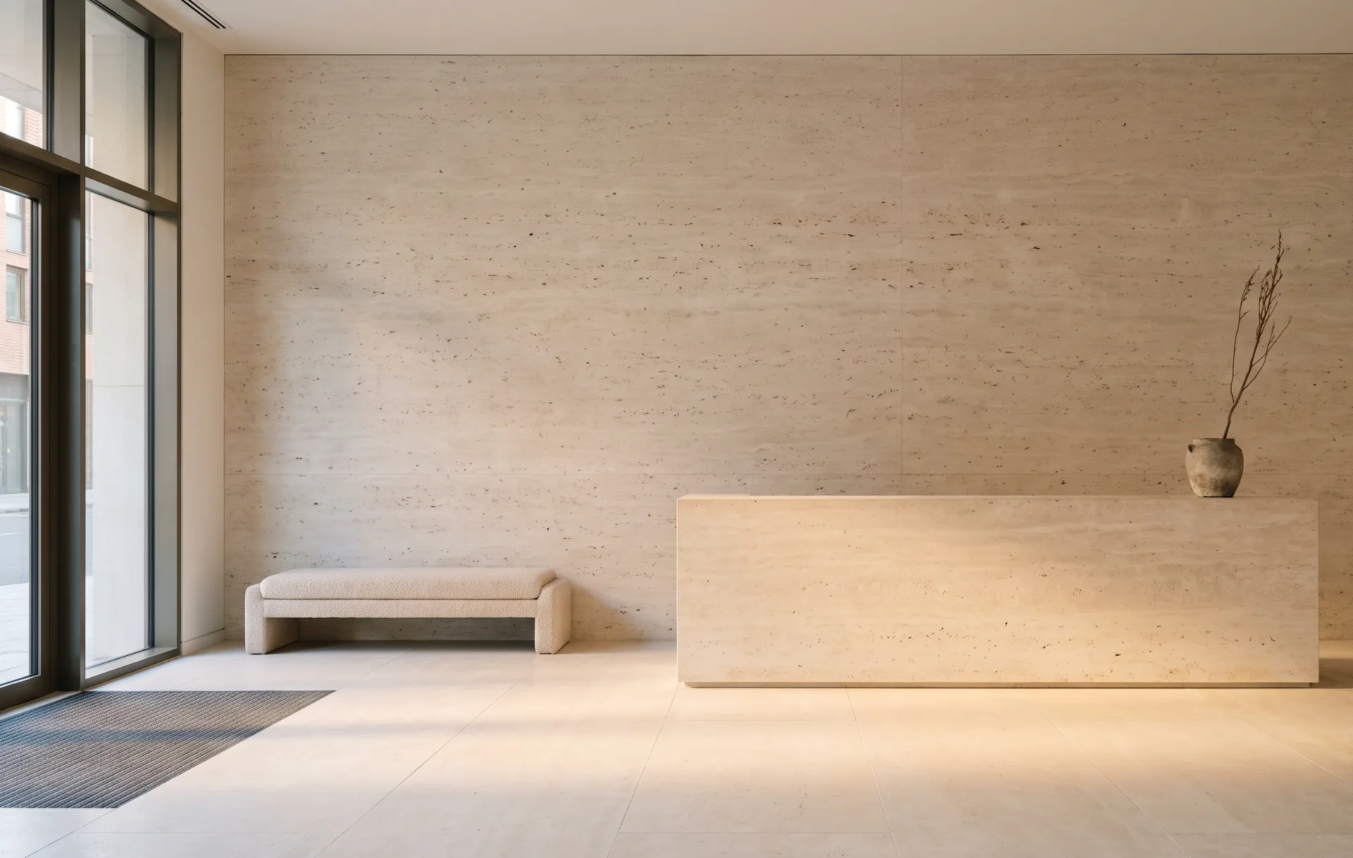 A quiet travertine reception hall with a stone counter, a bouclé bench and full-height glazing onto the street.