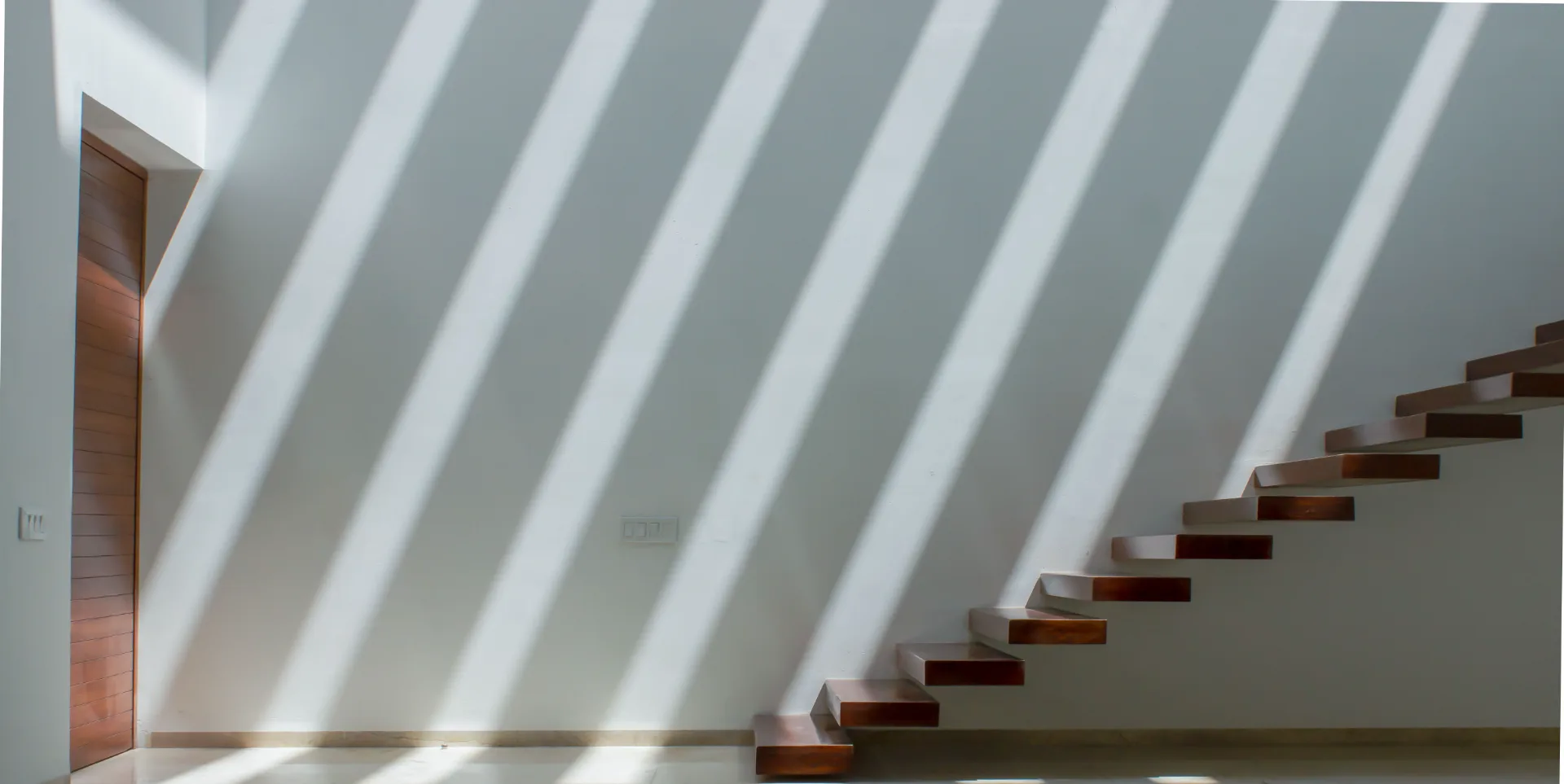 Sunlight falling in broad diagonal bands across a plastered wall, beside a flight of cantilevered timber treads.