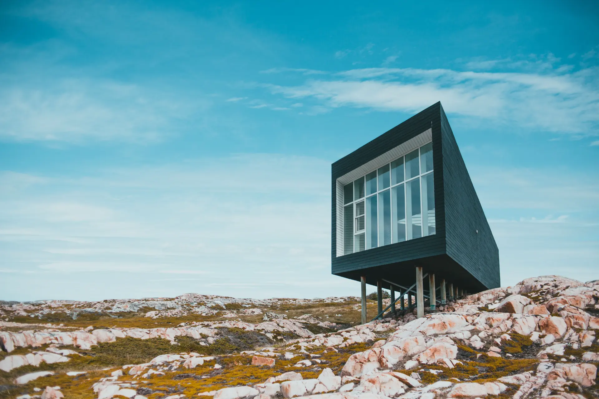 A dark timber house raised on slender columns above bare rock, standing alone against an open sky.